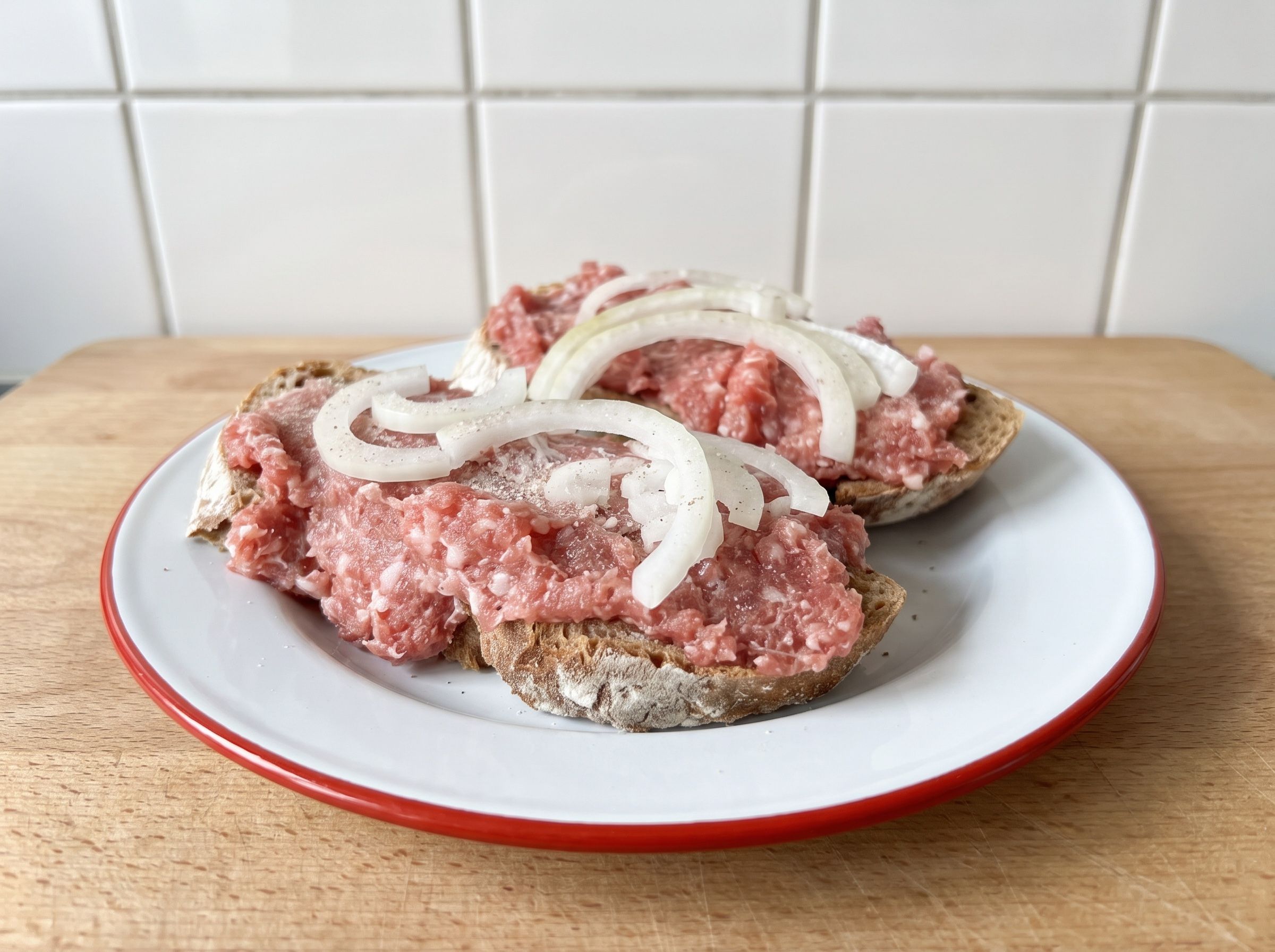 Zwei Scheiben Graubrot mit Mett und Zwiebelringen auf einem Emailleteller mit rotem Rand